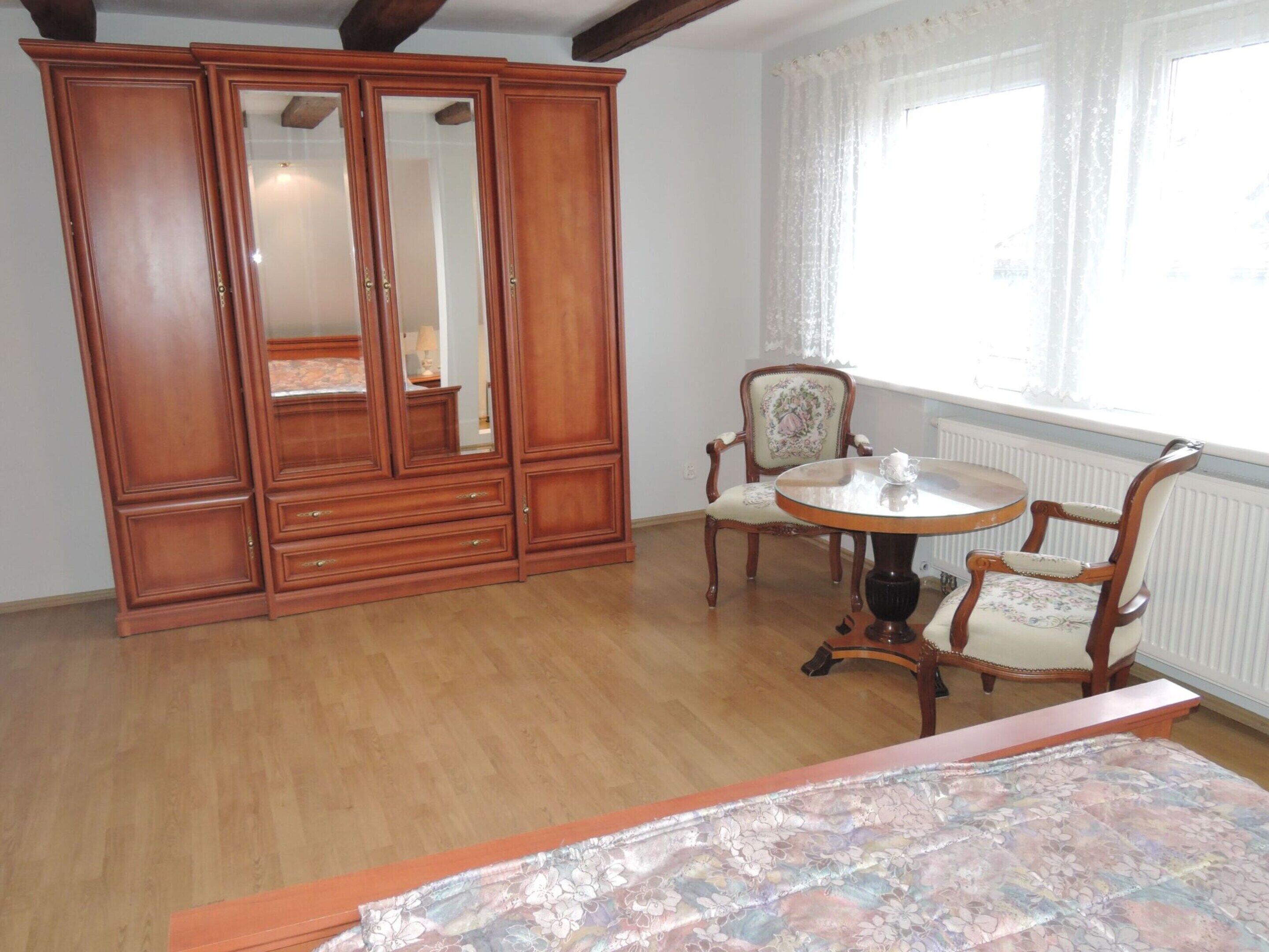 Une pièce comprenant une armoire en bois, une table ronde avec des chaises et un lit recouvert d’une couvre-lit à motifs floraux.