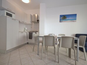 A modern kitchen with white cabinets, a microwave, and a gas stove. There is a dining area with a table and chairs, and a painting of a beach on the wall. - Residence Marco Polo, Lignano Sabbiadoro (Lignano Sabbiadoro)