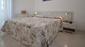 A bedroom with a bed, a nightstand, a lamp, a painting, and a window with curtains. - Residence Marco Polo, Lignano Sabbiadoro (Lignano Sabbiadoro)