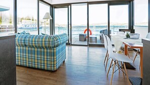 Ein modernes Interieur mit Sofa, Esstisch, Stühlen und großen Fenstern mit Blick auf ein Gewässer.