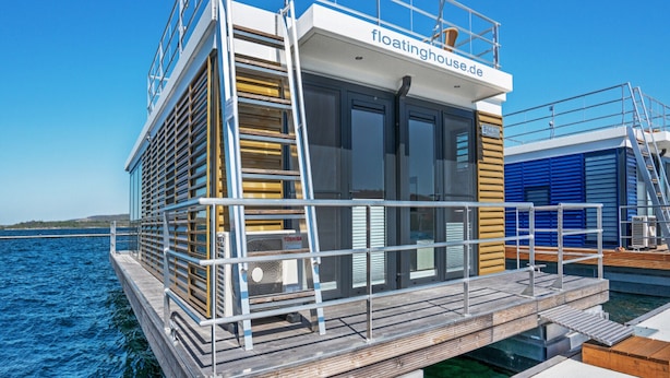 Ein schwimmendes Haus mit einer Terrasse, einer Leiter und einem Schild mit der Aufschrift 'floatinghouse.de'.