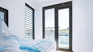 Ein Schlafzimmer mit Bett, einem Fenster mit Jalousien und einer Tür, die auf einen Balkon mit Blick auf das Wasser und Boote führt.
