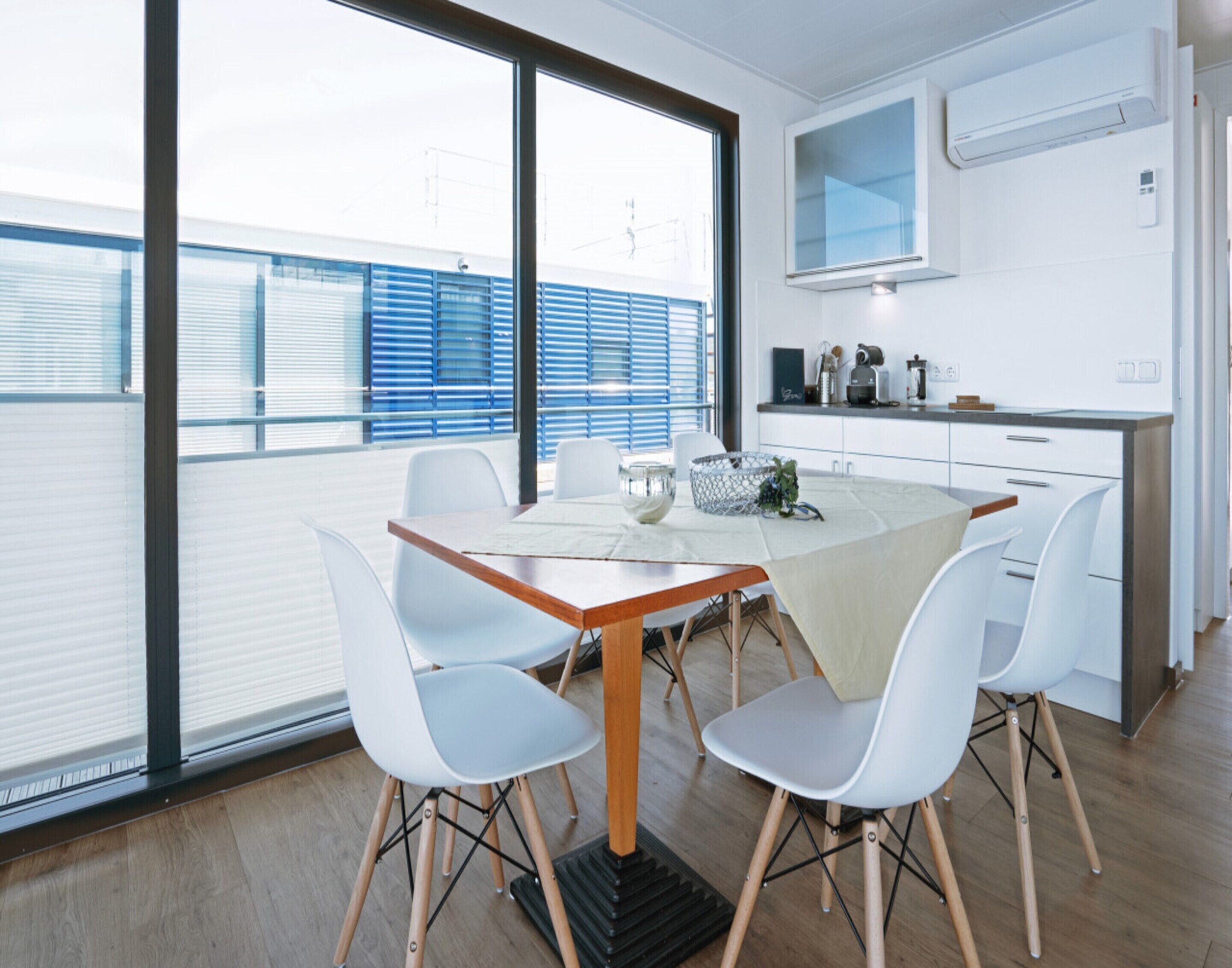 A modern dining area with a wooden table and white chairs, a kitchen with white cabinets and a microwave, and large windows with blinds.
