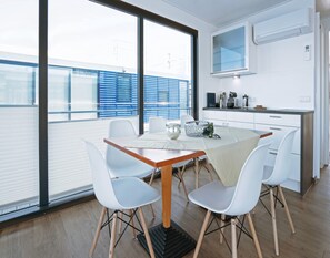 Un espace repas moderne avec une table en bois et des chaises blanches, une cuisine avec des meubles blancs et un four à micro-ondes, et de grandes fenêtres équipées de stores.