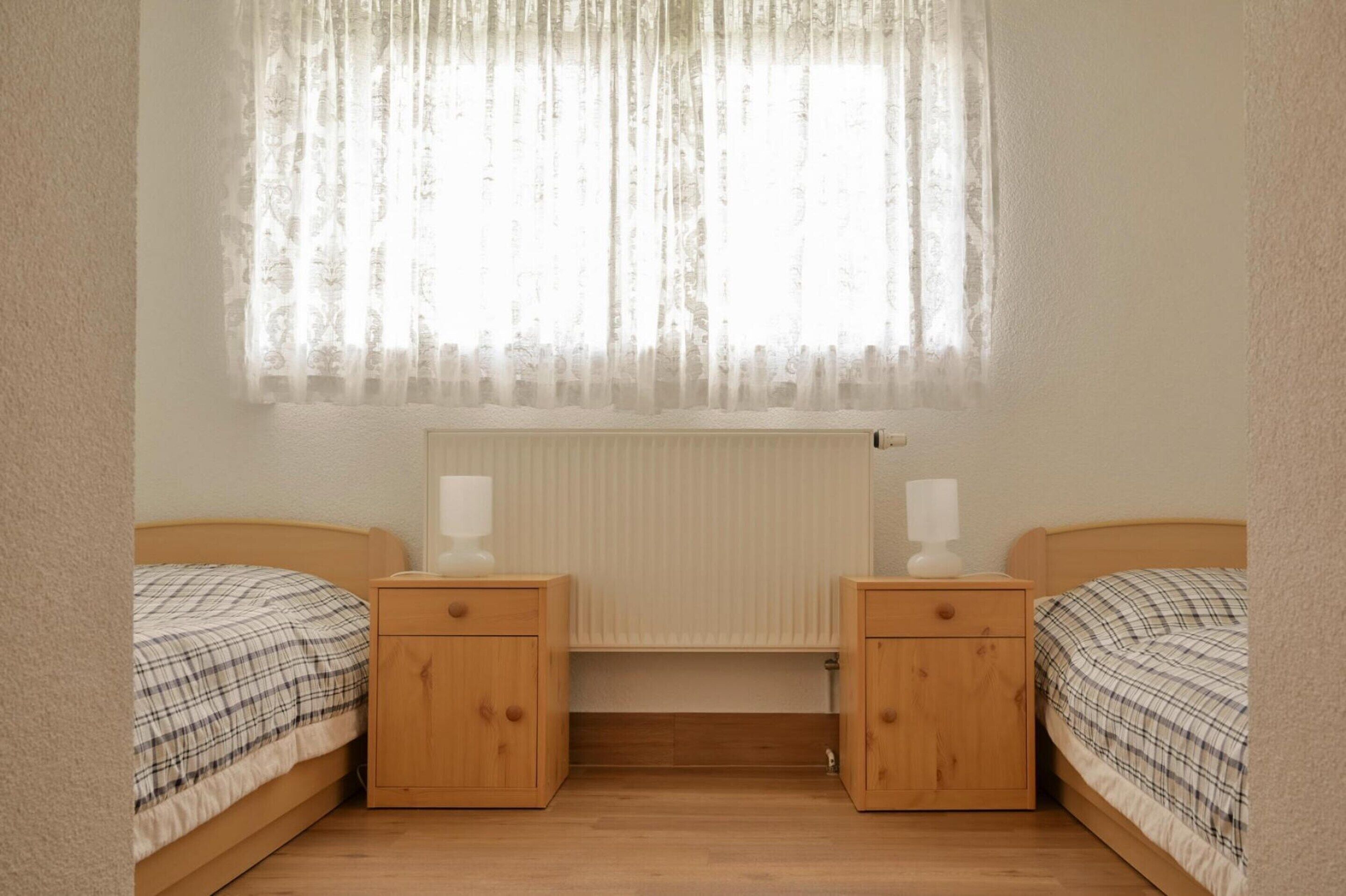 A bedroom with two beds, wooden nightstands, a radiator, and sheer curtains.