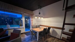 A dining area with a wooden table and chairs, a view of a garden through a sliding glass door, and a ceiling light fixture. - Ferienwohnung Tiefblick, Dornumersiel (Dornum)