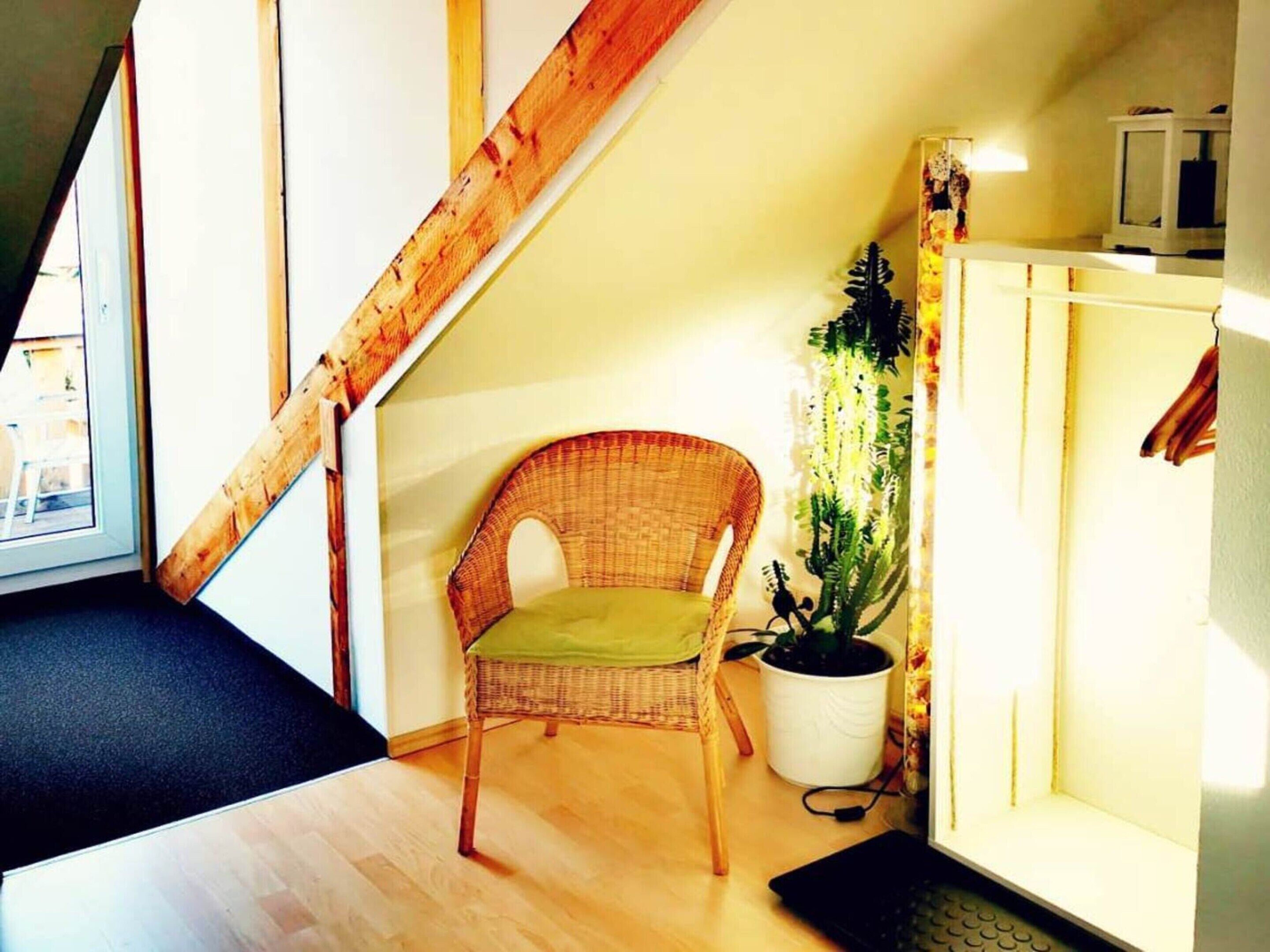 A room with a wooden staircase, a wicker chair with a green cushion, a potted plant, and a white cabinet with a television.