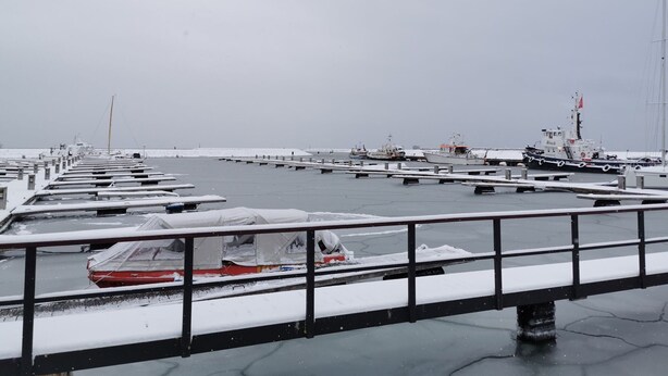 Una marina con barcos amarrados, cubiertos de nieve, y un puente al frente.