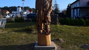 Una estatua de un hombre con abrigo y sombrero, sobre un pedestal en un área verde, con edificios y coches al fondo.