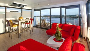 A modern living room with a red sofa, a dining area with chairs, and a large window overlooking a body of water. - Haus in Ribnitz Damgarten for 3 Personen (Ribnitz-Damgarten)