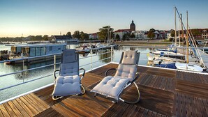 A wooden deck with two lounge chairs facing a marina with boats and a building in the background. - Haus in Ribnitz Damgarten for 3 Personen (Ribnitz-Damgarten)