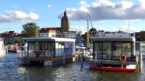 A riverfront with two houseboats docked, a prominent tower in the background, and a clear sky. - Haus in Ribnitz Damgarten for 3 Personen (Ribnitz-Damgarten)