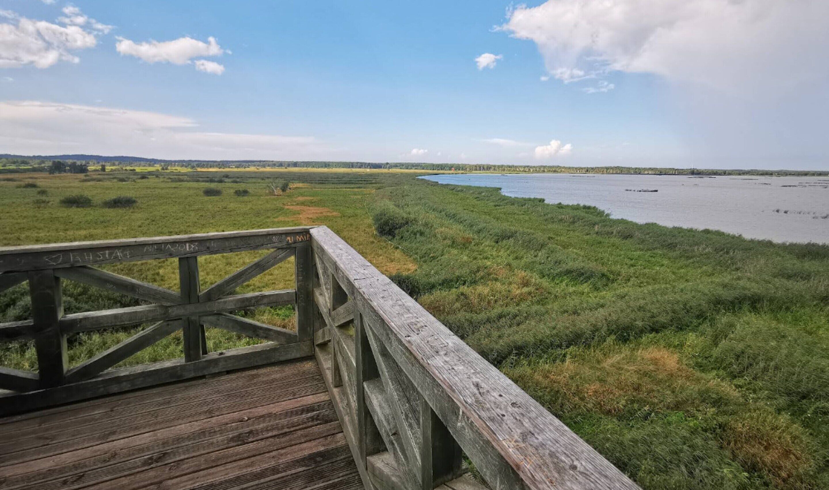 A wooden boardwalk leading to a river with grassy banks and distant hills.