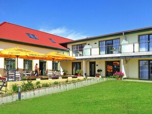 A two-story building with a red-tiled roof, outdoor seating area with yellow umbrellas, and a well-maintained lawn. - Appartements am Heidensee in Schwerin (Schwerin)