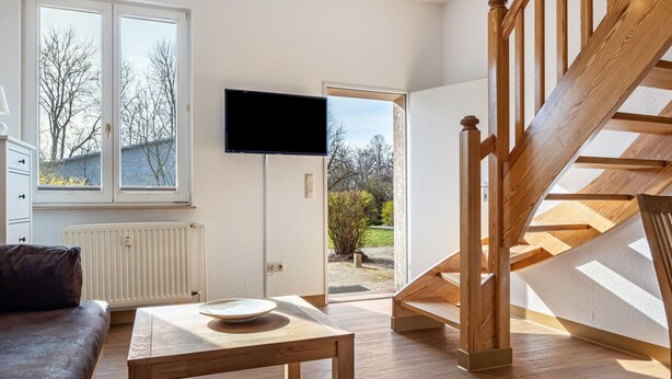 Una sala de estar moderna con una escalera de madera, un televisor de pantalla plana montado en la pared y vistas al exterior a través de amplios ventanales.
