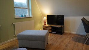 A room with a television on a wooden stand, a lamp, a window with a view, and a grey ottoman. - Ferienwohnung in Wyk-boldixum auf Foehr (Wyk auf Foehr)