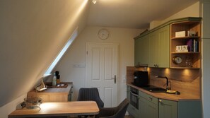 A compact kitchen with a dining area, a clock on the wall, and a shelf with books and a calendar. - Ferienwohnung in Wyk-boldixum auf Foehr (Wyk auf Foehr)
