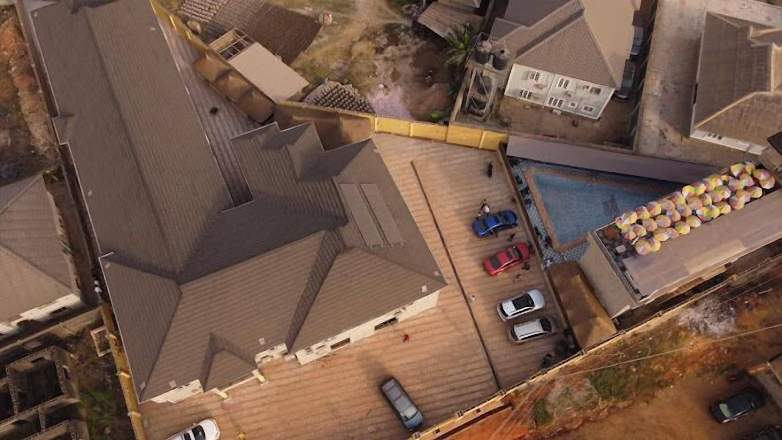 An aerial view of a residential area with multiple houses, a swimming pool, and several parked cars.