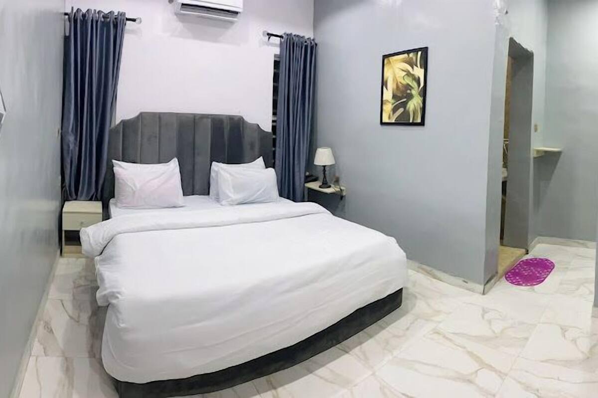 A hotel room with a large bed, two nightstands, a wall-mounted air conditioner, and a painting on the wall.