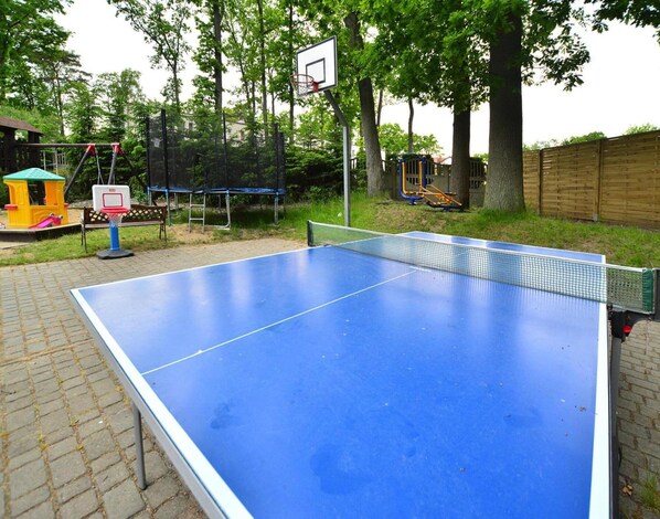 Une table de ping-pong installée à l’extérieur, avec un panier de basketball et des jeux pour enfants en arrière-plan.