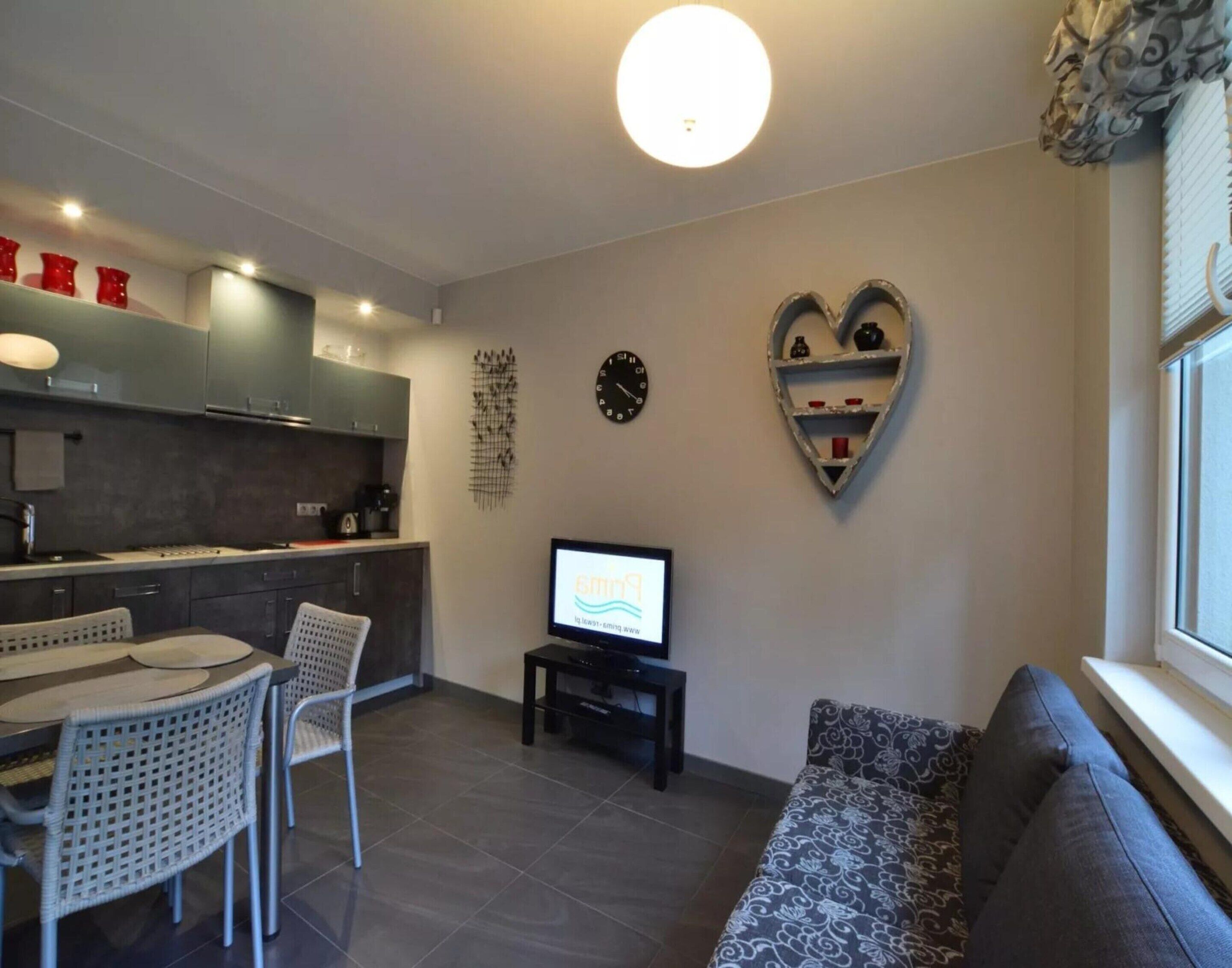 Een moderne woonkamer met een bank, een eettafel met stoelen, een televisie en een keukenhoek met kasten en een klok.