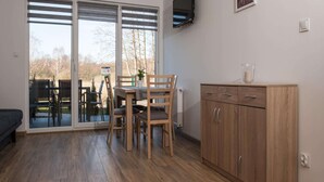 A modern living room with a wooden dining table, chairs, and a cabinet. Large windows with blinds provide a view of the outdoors. - Comfortable Ground Floor Apartment, Summer Pool (Postomino)