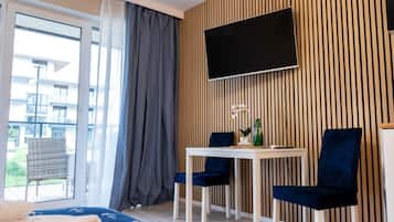 A modern hotel room with a large window, a flat-screen TV mounted on the wall, a small table with two chairs, and a bed with a blue blanket.