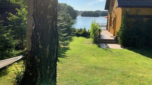 Une vue sur un lac, avec un hamac suspendu à un arbre, un sentier menant à un bâtiment et une végétation luxuriante.