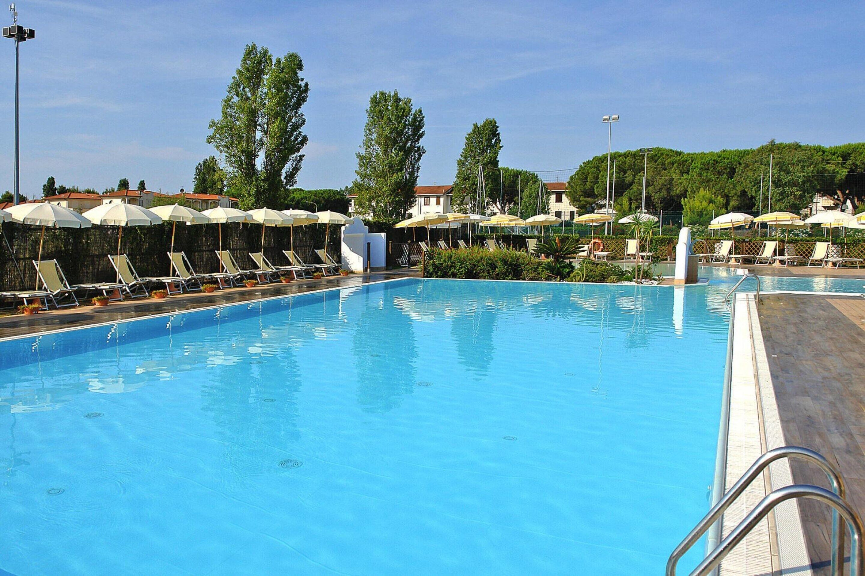 A swimming pool with sun loungers and umbrellas, surrounded by greenery and buildings.
