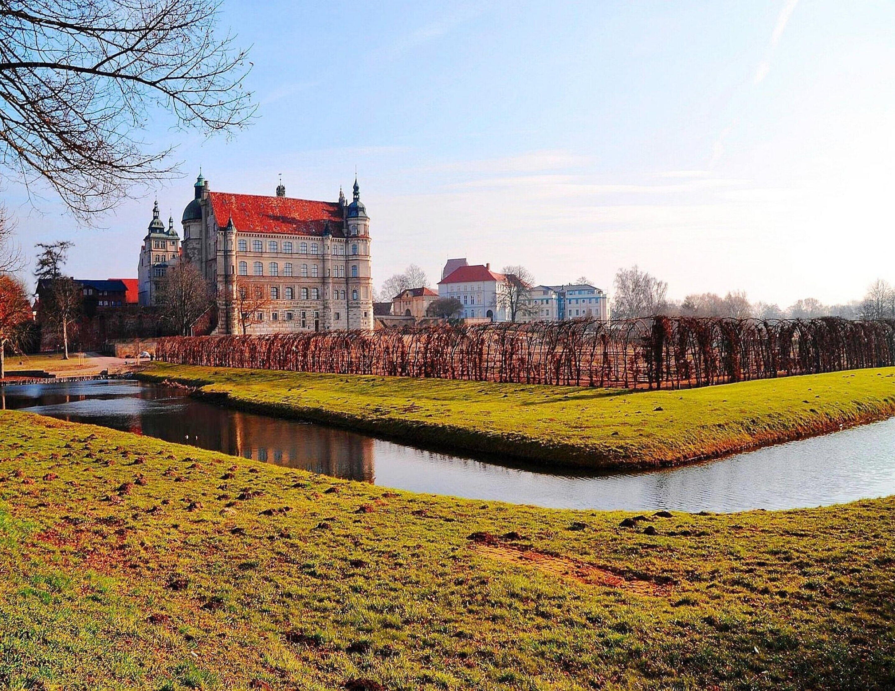 Ein historisches Gebäude mit rotem Dach und klassischer Architektur, ein Kanal und eine begrünte Fläche mit Bäumen.