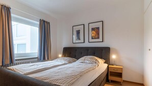 A bedroom with a double bed, a nightstand, two framed pictures, and a window with curtains. - Ferienwohnung, St. Peter - Ording (St. Peter-Ording)