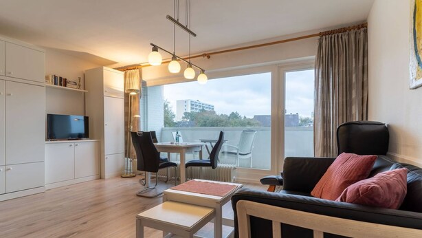 A modern living room with a sofa, a coffee table, and a dining area with chairs and a table. There is a TV on a cabinet, and a balcony with a view of buildings. - Ferienwohnung, St. Peter - Ording (St. Peter-Ording)
