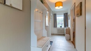 A hallway with a white bench, a radiator, a window with curtains, and a hanging lamp. - Ferienwohnung, St. Peter - Ording (St. Peter-Ording)
