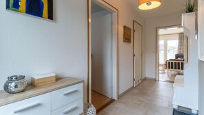 A hallway with a wooden floor, white walls, and a door leading to a room. There is a small table with a vase and a box on it, and a painting on the wall. - Ferienwohnung, St. Peter - Ording (St. Peter-Ording)