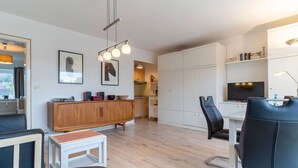 A modern living room with a wooden cabinet, a white desk, a black leather chair, and a small white table. - Ferienwohnung, St. Peter - Ording (St. Peter-Ording)