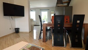 Uma sala de estar moderna com uma televisão de tela plana fixada na parede, um espaço de refeições com uma mesa e cadeiras de madeira, e um corredor que dá para outro cômodo.