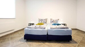 Uma cama de casal com roupa de cama estampada, duas mesas de cabeceira com abajures e uma janela grande em uma das paredes.