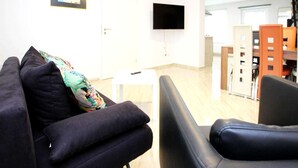 Uma sala de estar com sofá, uma poltrona, uma mesinha e uma televisão fixada na parede.