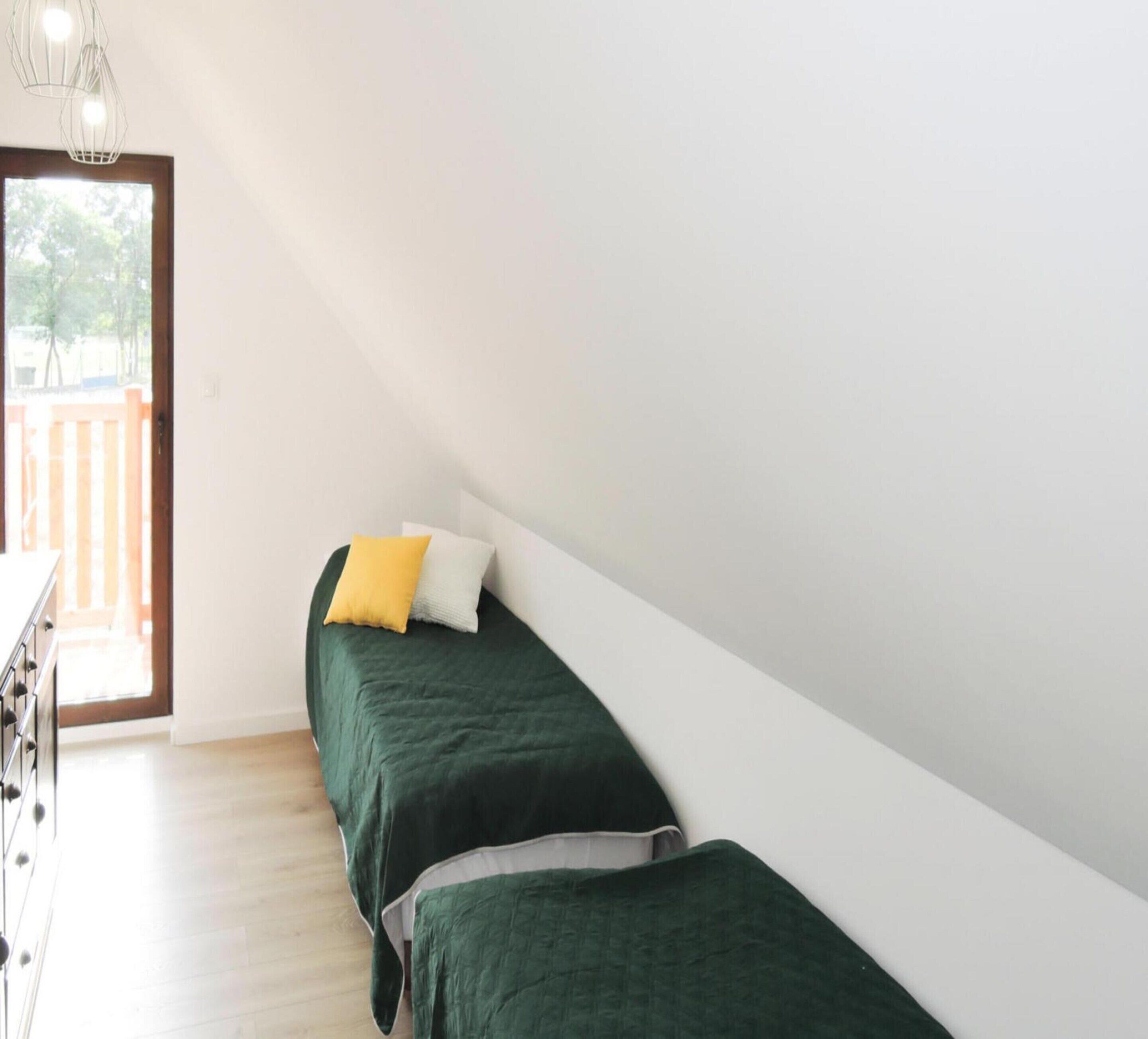 Una habitación pequeña con cama individual, una colcha verde, un almohadón amarillo y un almohadón blanco. Hay una puerta de madera que da a un espacio al aire libre.