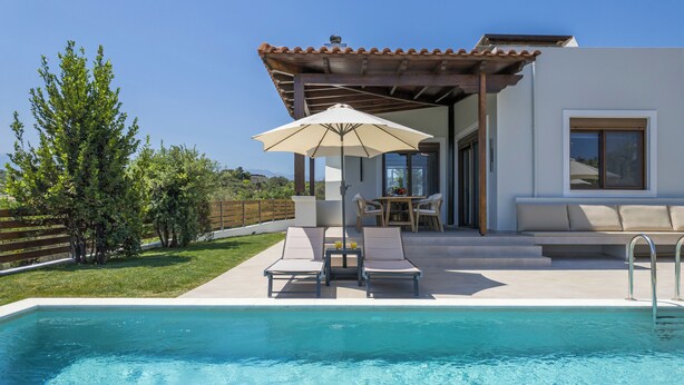 A pool area with lounge chairs and an umbrella, a patio with a table and chairs, and a view of the surrounding landscape. - Villa Wisdom (Chania)