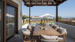 A wooden table with cushioned chairs, a glass door, and a view of a swimming pool and mountains. - Villa Wisdom (Chania)