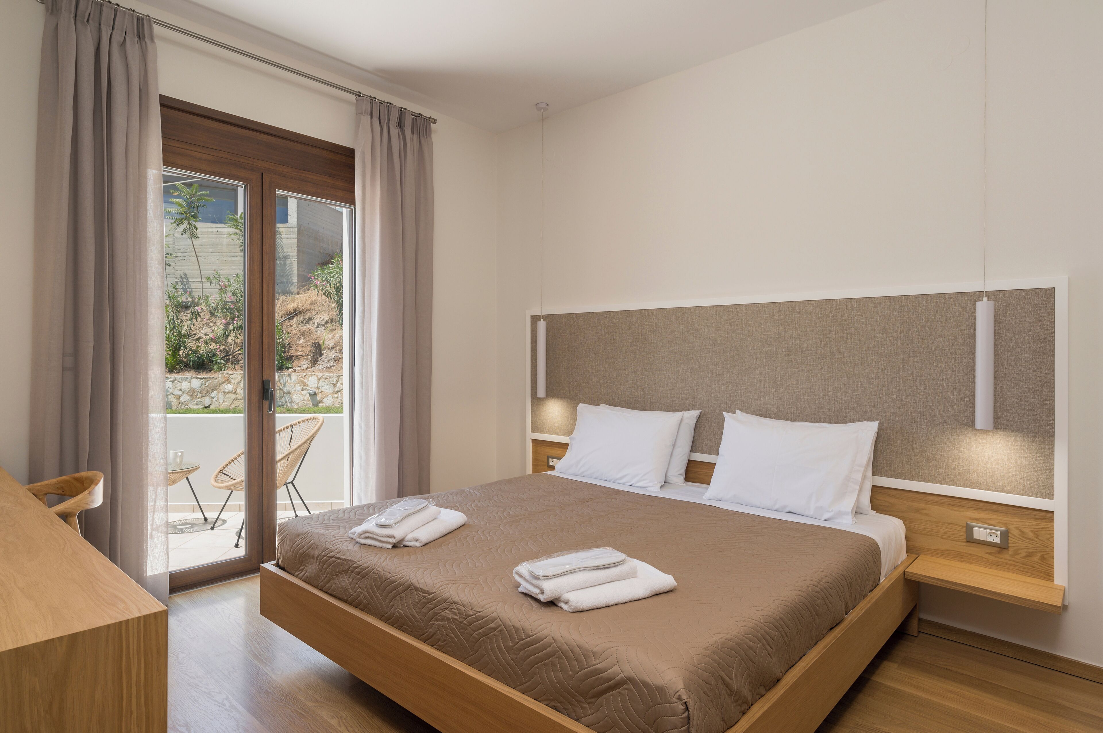 A modern hotel room with a large bed, a wooden nightstand, a desk, and a sliding glass door leading to an outdoor area.
