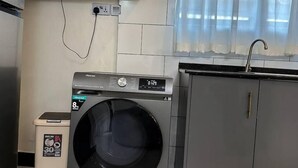 A compact laundry room with a front-loading washing machine, a small trash bin, and a grey cabinet. - LivLuxe Apartments 6 (Kampala)