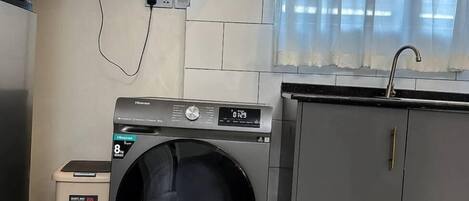 A compact laundry room with a front-loading washing machine, a small trash bin, and a grey cabinet.