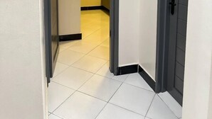 A hallway with a tiled floor, a door with a black handle, and a light switch on the wall. - LivLuxe Apartments 6 (Kampala)