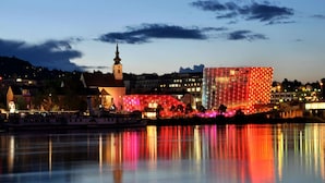 A waterfront cityscape with illuminated buildings, a church steeple, and a calm river reflecting the lights. - Appartements Nicht Weit von der Donau (Linz)