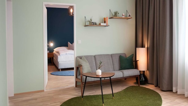 A living room with a sofa, a small table, a lamp, and a green rug. There is a bed in the background and a door leading to another room. - Appartements Nicht Weit von der Donau (Linz)
