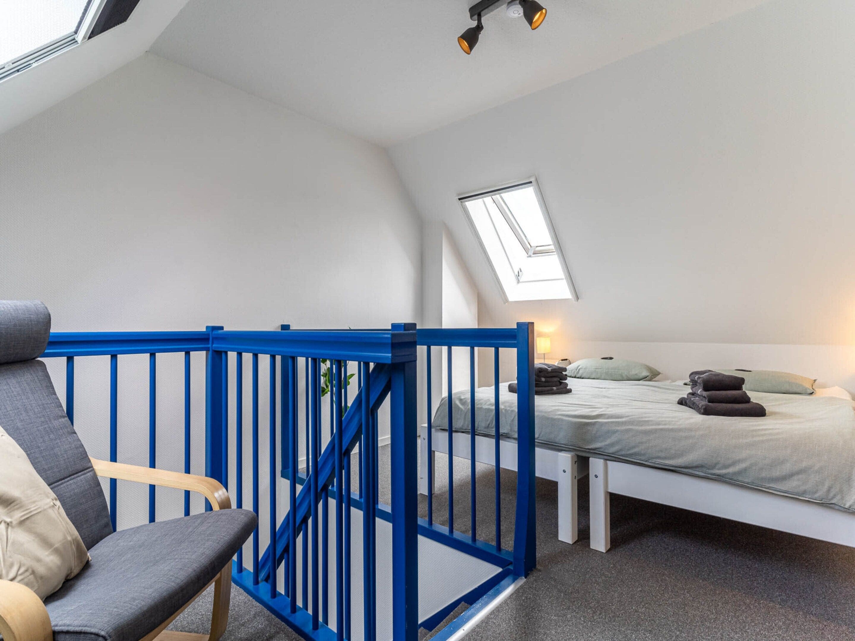 Une chambre avec un puits de lumière, un lit, une chaise et une rambarde bleue.