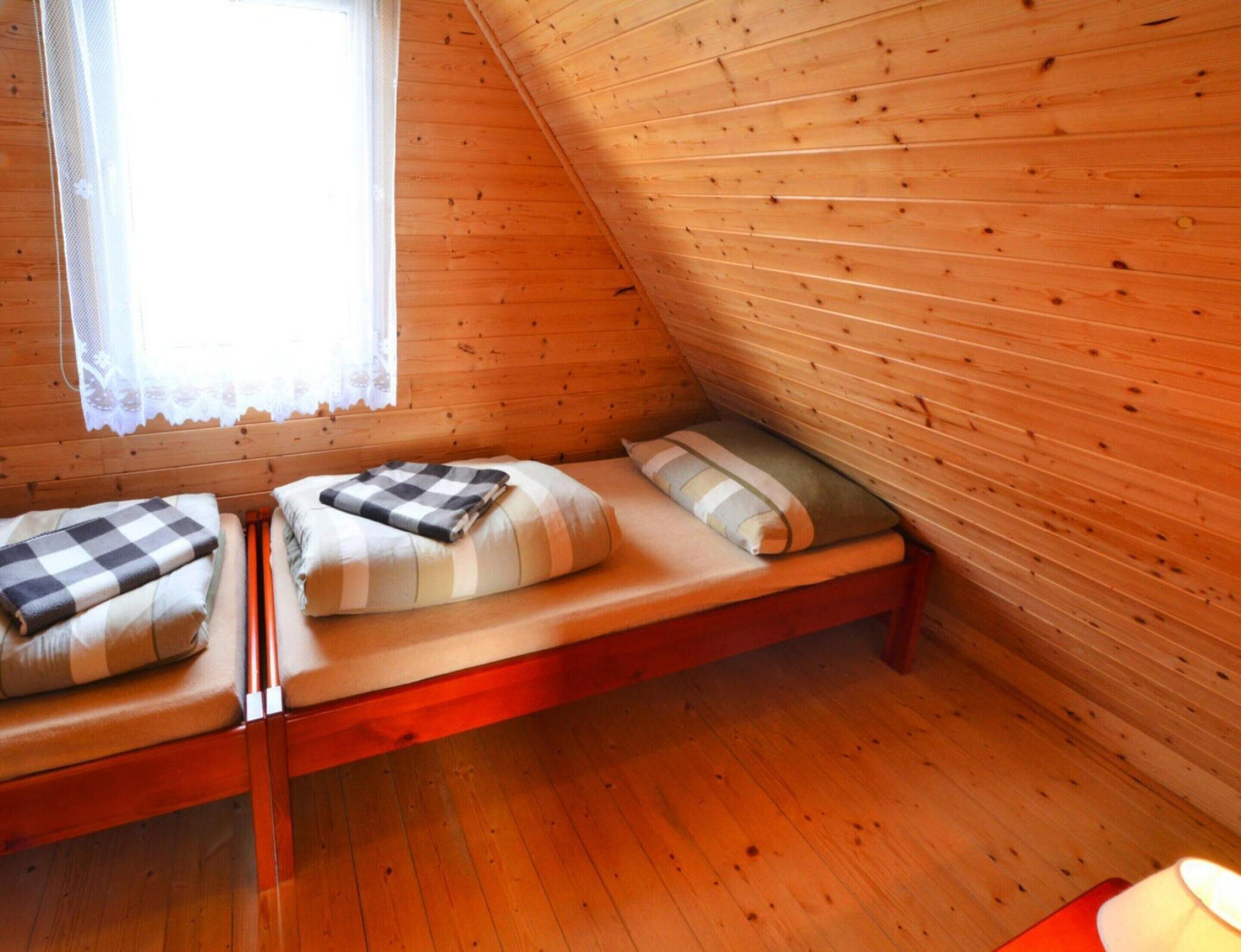 A small, cozy room with wooden walls and a sloped ceiling. There are two beds with pillows and blankets, a window with lace curtains, and a wooden floor.