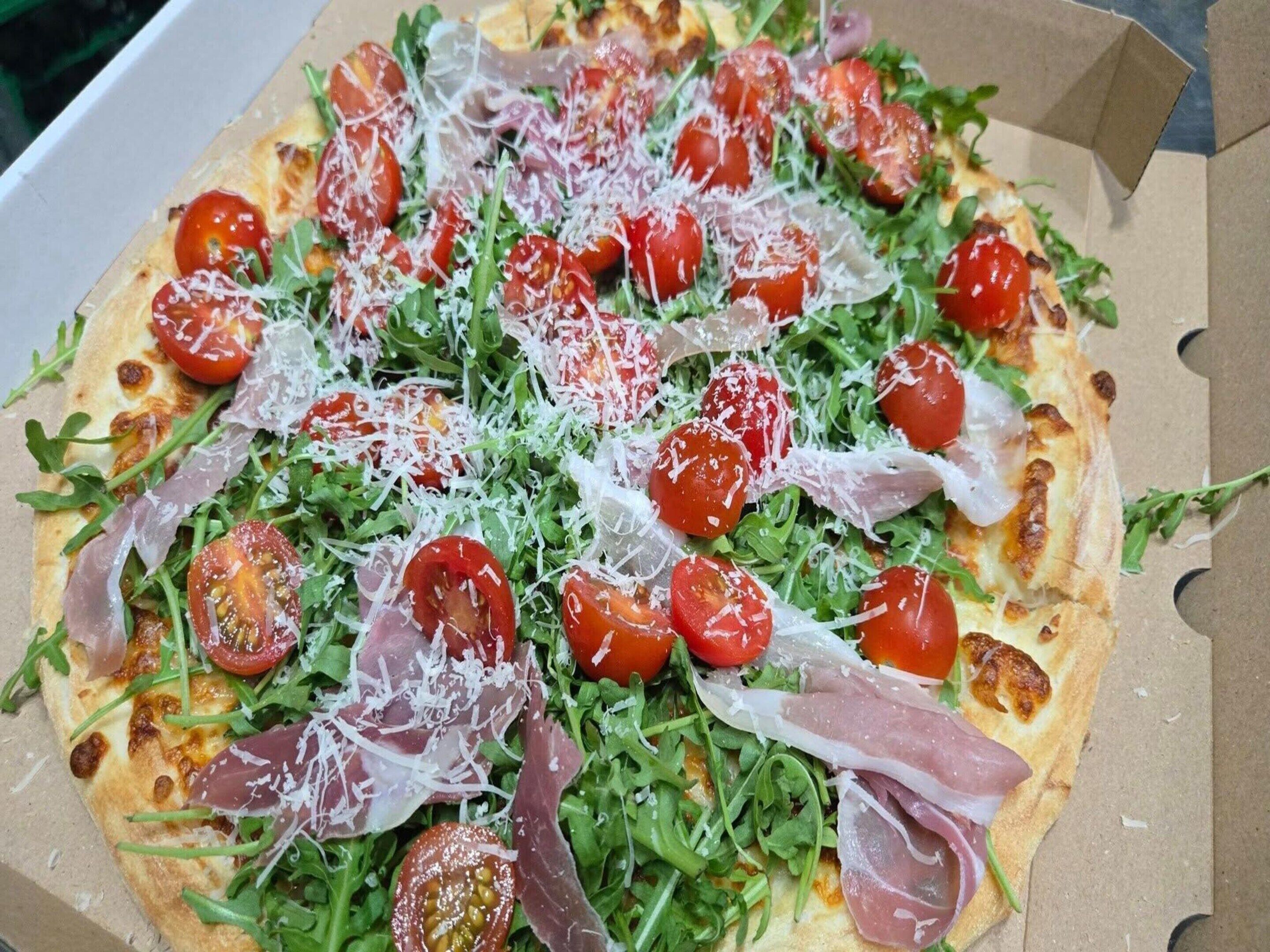 A pizza with arugula, cherry tomatoes, and prosciutto in a cardboard box.